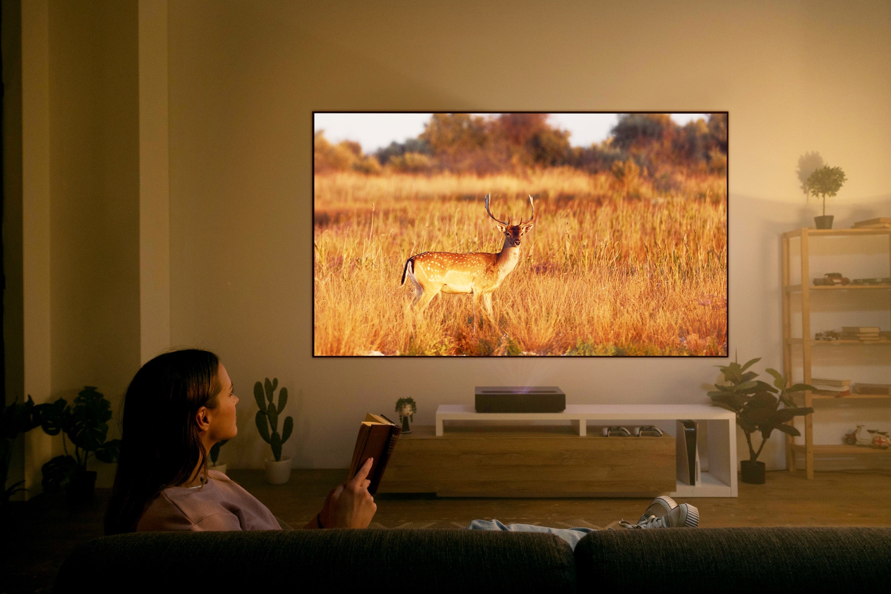 A person relaxes on a couch with a book, watching a large TV screen showing a deer in a sunlit field. The ambiance is enhanced by the FORMOVIE Theater 4K UHD UST Laser Projector, delivering stunning clarity and vibrant colors with Dolby Vision, while plants and decor add to the cozy feel.