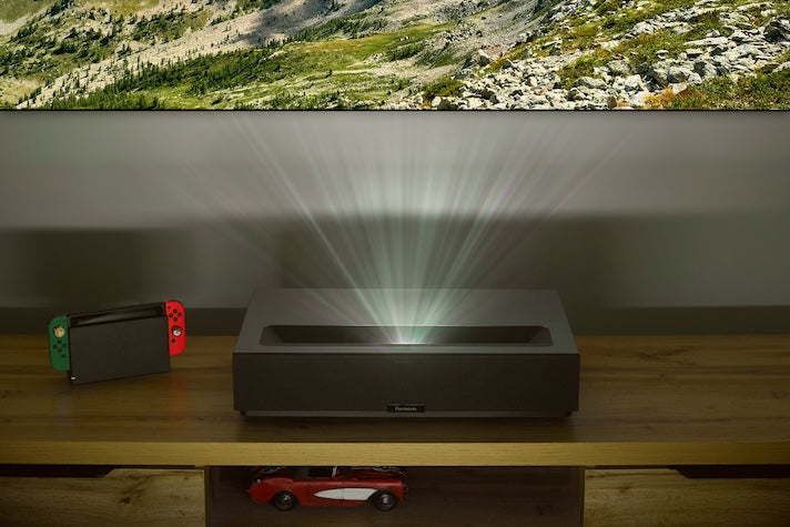A black FORMOVIE Formovie Theater 4K UHD UST Laser Projector on a wooden table displays a mountain scene in crystal-clear 4K. Nearby, a gaming console with red and green controllers sits beside a red toy car on the lower shelf.