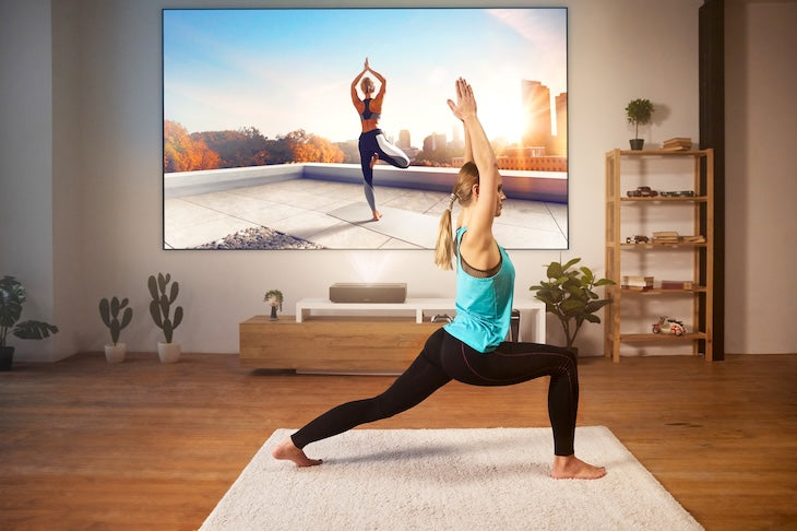 In a serene living room with wooden floors, plants, and a shelf, someone practices yoga while their FORMOVIE Formovie Theater 4K UHD UST Laser Projector displays a rooftop yoga routine in stunning clarity.