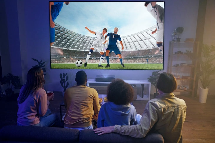 Four friends relax on a couch in a cozy living room, enjoying a soccer match in brilliant clarity with their FORMOVIE Theater 4K UHD UST Laser Projector. Players in white and blue uniforms are vivid under Dolby Vision, while soft lighting and plants enhance the inviting atmosphere.