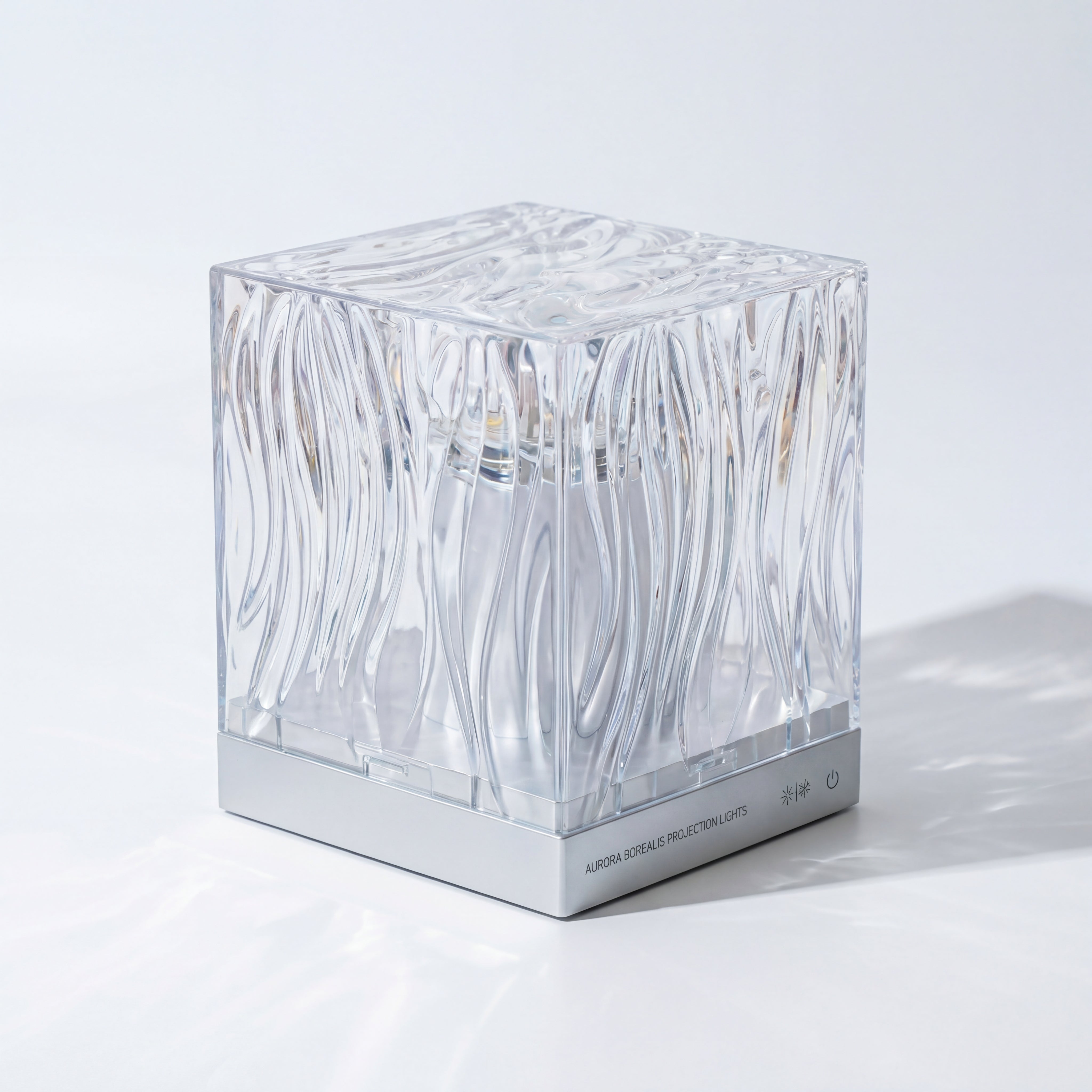 The Projection Room’s Aurora Projector Lamp is a transparent, textured cube with wave-like patterns and a silver power button at the base, creating mesmerizing ambient lighting for any space.
