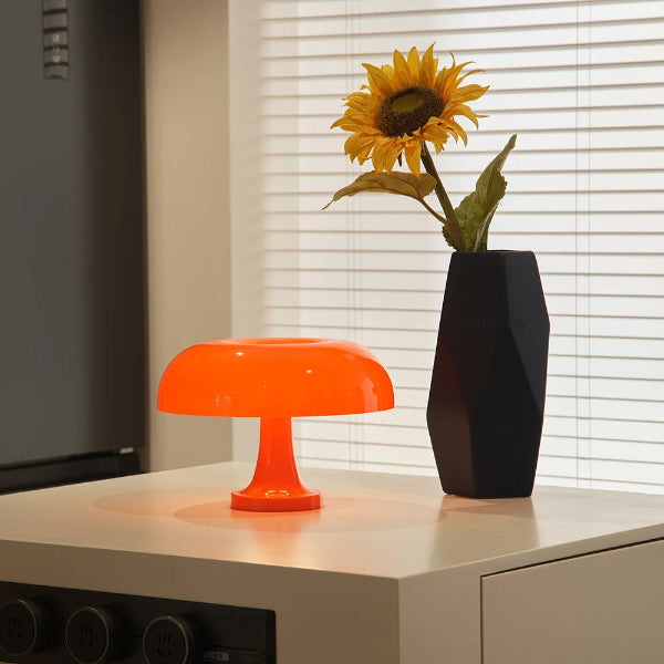 A Modern Danish Mushroom Table Lamp LED Bedside Lamp by The Projection Room sits on a white table beside a tall black vase with a sunflower, with white blinds in the background.