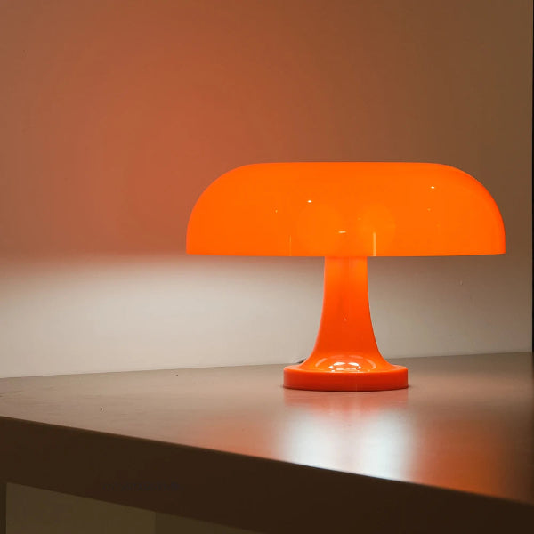 The Projection Rooms Modern Danish Mushroom Table Lamp LED Bedside Lamp adds a warm glow to a gray surface, its orange, mushroom-shaped design and energy-efficient dimmable LED lighting perfect for minimalistic spaces.