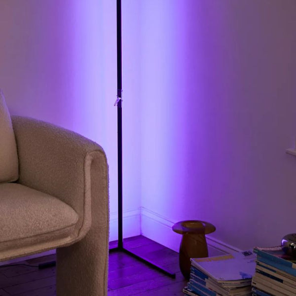 A cozy room corner with a plush, light-colored chair and the SMART LED Stand by The Projection Room casts a soft purple glow on the walls. Nearby, a small wooden stool holds books on the wooden floor.