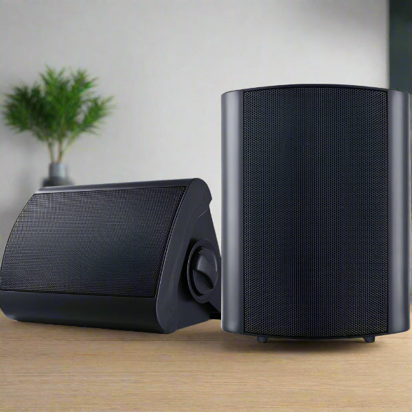 Two The Projection Room 2-Way In Wall Speakers, designed for 150W immersive sound, are on a wooden surface. One is upright and the other slightly tilted back. A blurred potted plant sits in the background against a plain wall.