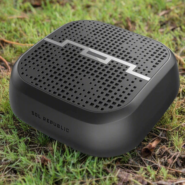 A black, square-shaped SOL Republic Punk Wireless Pocket Speaker by The Projection Room with a perforated grill sits on the grass. It features a silver emblem on top and a subtle side logo, providing impressive Bluetooth range for outdoor adventures.