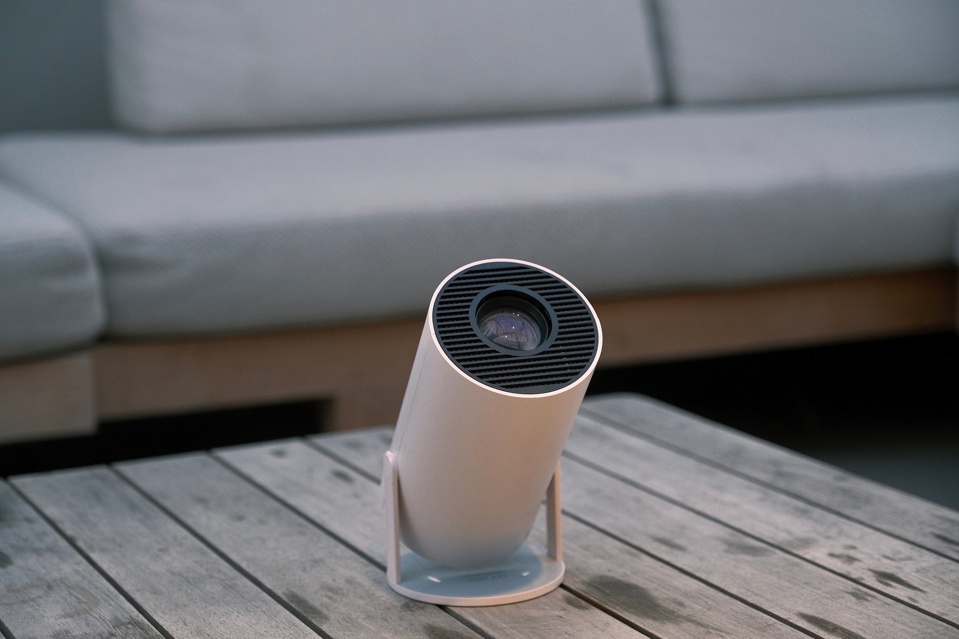 A modern white projector sits on a wooden table in front of a light-colored sofa with cushions, creating a cozy indoor setting.