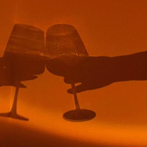 The Projection Rooms Sunset Glow LED Lamp features silhouettes of two clinking wine glasses against a warm orange backdrop, enhanced by modern lighting. A hand holds one glass, adding elegance.