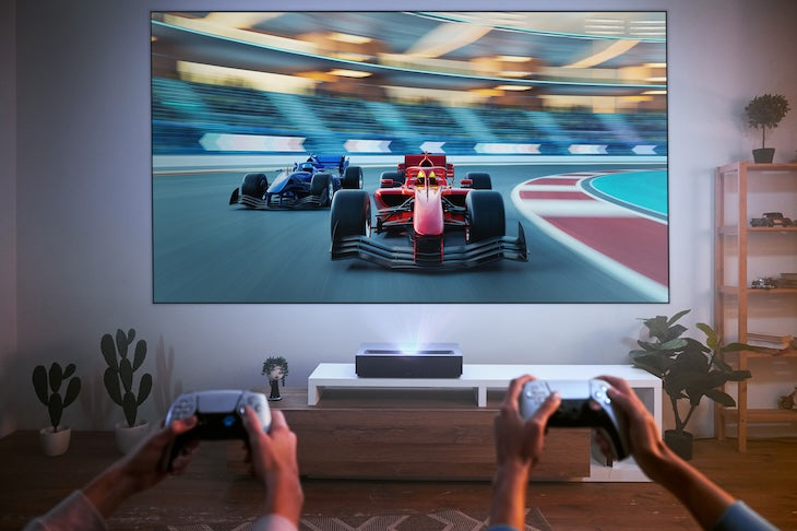 In a modern living room, two people holding controllers play a racing game on a large screen. The fast-paced action is projected by the FORMOVIE Theater 4K UHD UST Laser Projector on a low wooden table, with potted plants adding greenery to the scene.