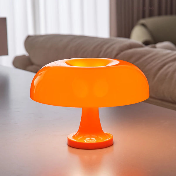 The Projection Room’s Modern Danish Mushroom Table Lamp, featuring dimmable LED lighting and a bright orange design, sits on a white table in a modern living room with neutral furniture and soft natural light.