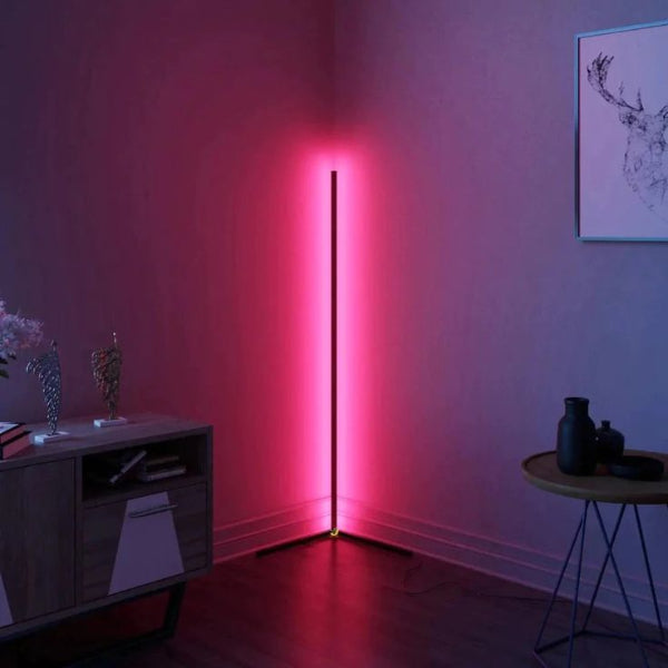 A minimalist room showcases The Projection Rooms SMART LED Stand, a tall, energy-efficient corner lamp emitting soft pink light with voice and app controls. A wooden sideboard with decorative items sits to the left, a round table with dark objects to the right, and framed artwork adorns the wall.
