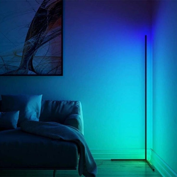 In a modern, dimly-lit room, a grey couch with a draped blanket sits beside The Projection Rooms SMART LED Stand. This tall, thin floor lamp emits energy-efficient LED light in blue and green hues. An abstract painting on the wall subtly harmonizes with the lamps RGB color options.