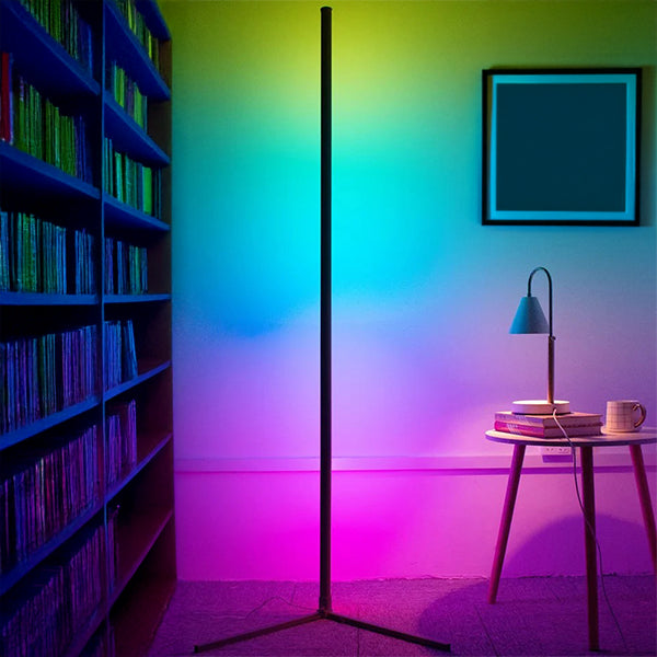 A modern SMART LED Stand by The Projection Room, with energy-efficient technology, casts customizable blue, green, and pink light on the wall. Next to it sits a round table with books, a mug, and a small lamp. Shelves of books line the left side while a framed picture hangs on the wall.