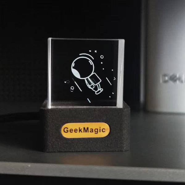 A small glass cube engraved with an astronaut floats in space and is displayed on a black stand, labeled The Projection Room. The customizable 3D Icon Desk Light Decor has a backdrop showing part of a shelf and a partially visible computer tower.