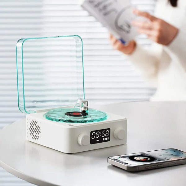 A Vinyl Speaker Lamp from The Projection Room with a water ripple RGB light and transparent cover sits on a white table, producing high-quality sound. An iPhone shows the interface nearby while a book-holding person in a white sweater appears blurred in the background.
